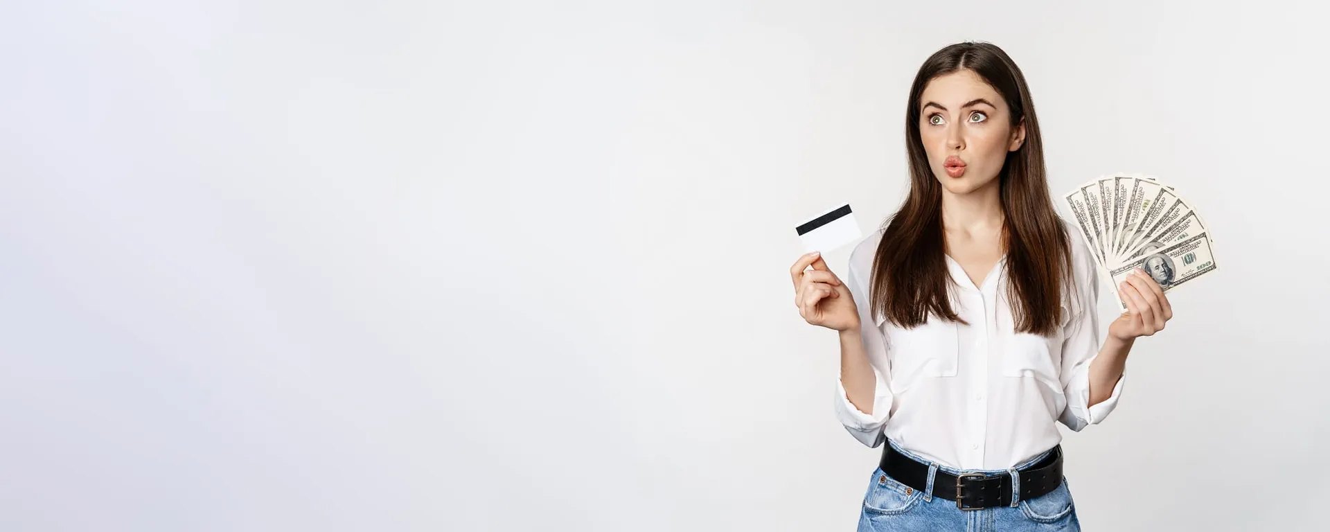 Surprised young woman holding cash and a card against a light background